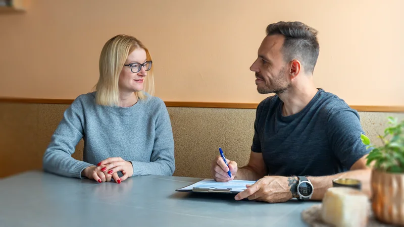 Sascha im Gespräch mit einer Klientin am Küchentisch, Notizen auf dem Klemmbrett